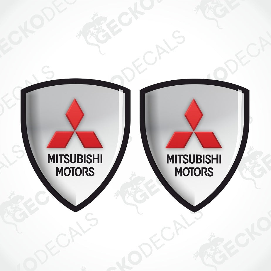 Two identical shield-shaped Mitsubishi Motors decals on a light gray background with diagonal watermark text. Each shield has a black border, a silver gradient center, the red三菱 logo, and black text reading 'MITSUBISHI MOTORS'.