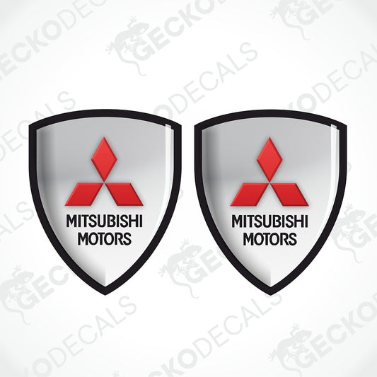Two identical shield-shaped Mitsubishi Motors decals on a light gray background with diagonal watermark text. Each shield has a black border, a silver gradient center, the red三菱 logo, and black text reading 'MITSUBISHI MOTORS'.
