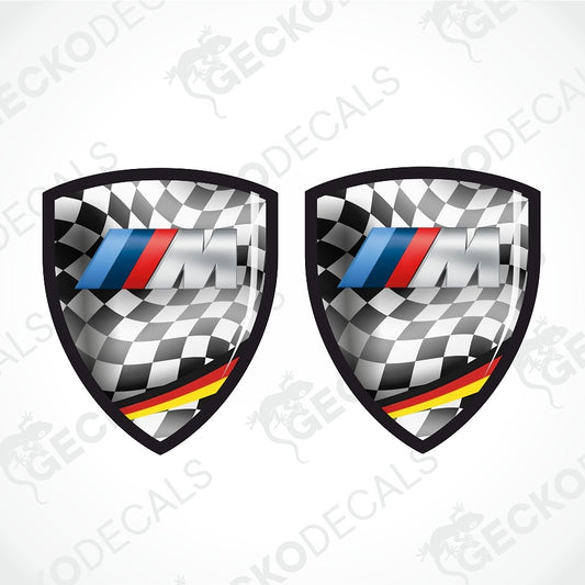 Two identical shield-shaped decals with a black-and-white checkered flag pattern, a blue, red, and white striped M logo, and a yellow, red, and black stripe near the bottom, shown on a light gray background with repeated watermark text.