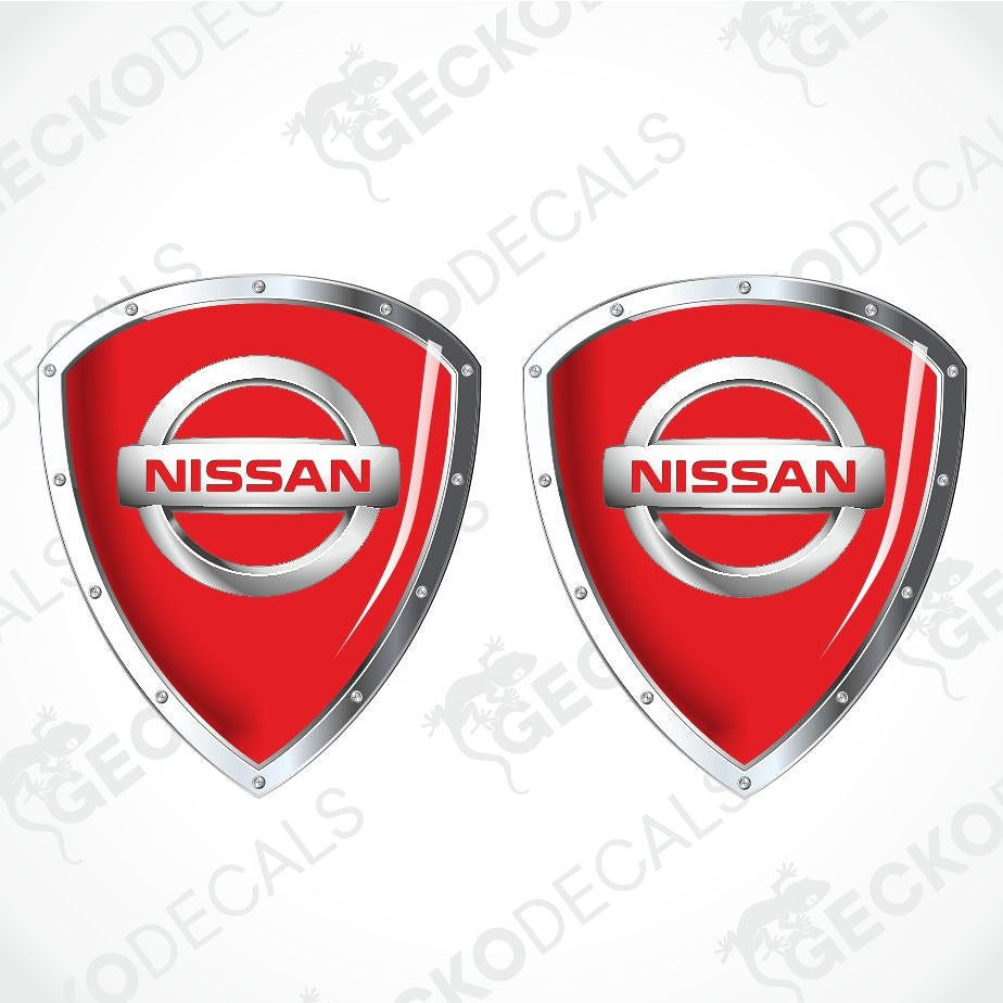 Two red Nissan shield-shaped decals with silver metallic borders and the Nissan logo in the center, shown on a light gray background with faint watermark text.