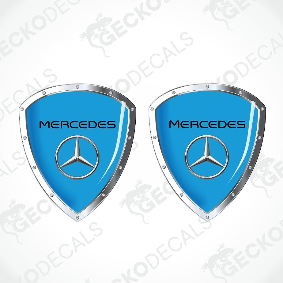 Two identical Mercedes-Benz shield-shaped decals on a light gray background with diagonal watermarks. Each decal has a bright blue center, a silver metallic border, and the Mercedes star logo in the middle with the word 'Mercedes' above it.