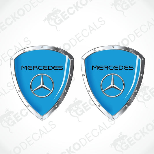 Two identical Mercedes-Benz shield-shaped decals on a light gray background with diagonal watermarks. Each decal has a bright blue center, a silver metallic border, and the Mercedes star logo in the middle with the word 'Mercedes' above it.