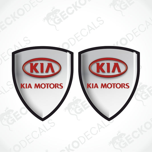 Two identical shield-shaped Kia Motors decals on a light gray background with diagonal watermarks. Each shield has a black border, a white center, a red oval Kia logo, and red 'KIA MOTORS' text.