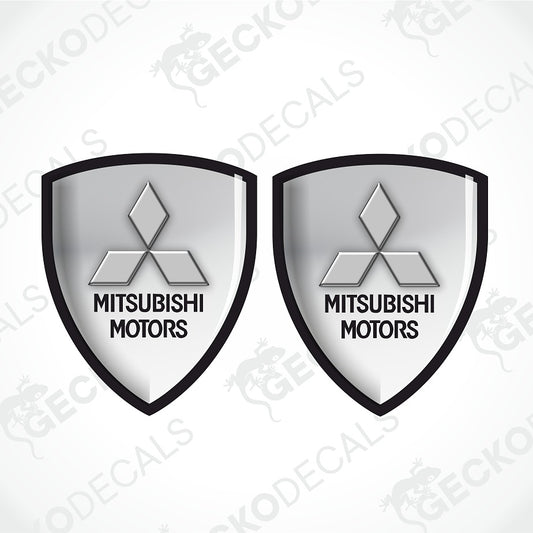 Two identical Mitsubishi Motors shield-shaped decals on a light gray background with diagonal watermarks. Each shield is silver-gray with a black border and the Mitsubishi logo and text.