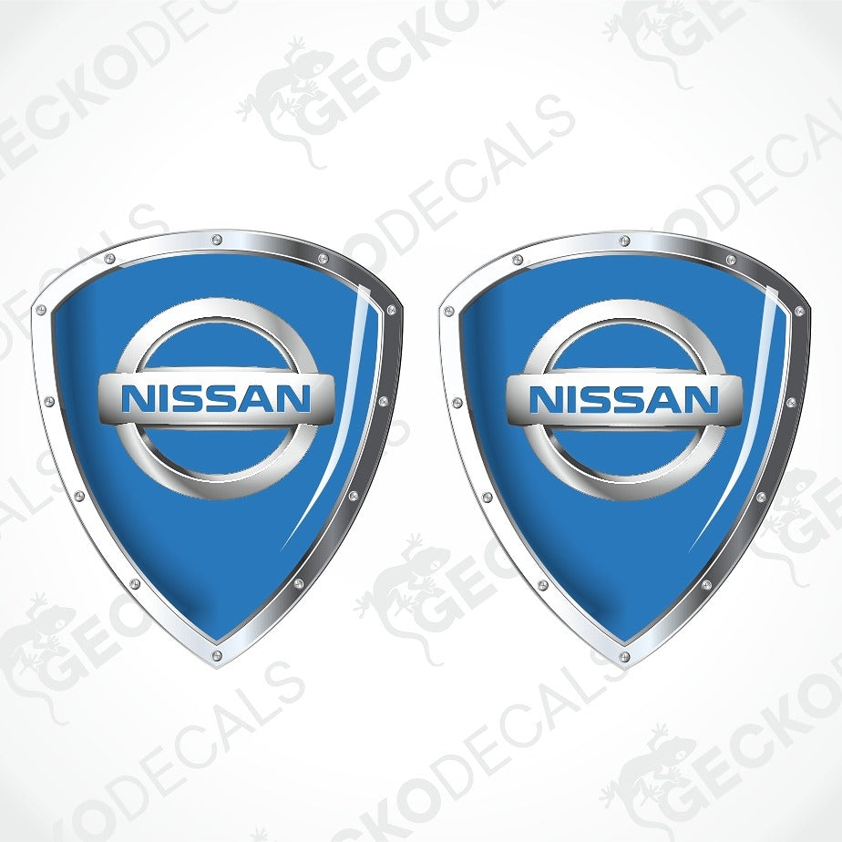 Two blue Nissan shield-shaped decals with silver metallic borders and the Nissan logo in the center, shown on a light gray background with faint watermark text.