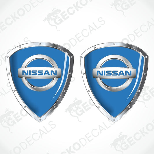 Two blue Nissan shield-shaped decals with silver metallic borders and the Nissan logo in the center, shown on a light gray background with faint watermark text.
