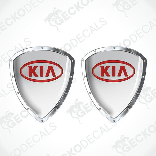Two identical glossy shield-shaped car decals with silver borders and the red Kia logo in the center, shown on a light gray background with faint watermark text.