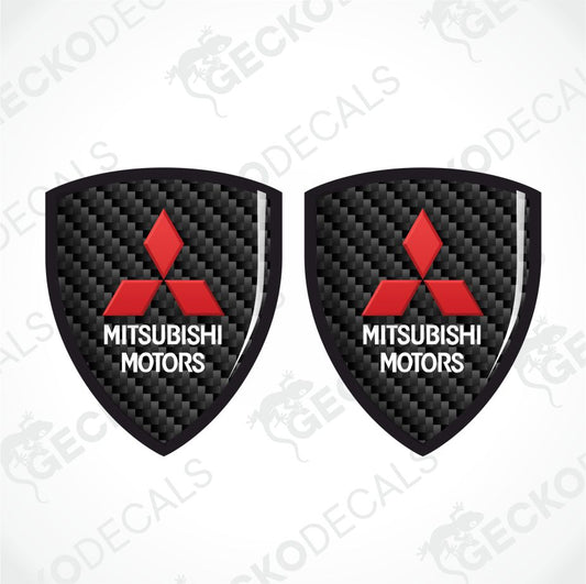 Two identical shield-shaped decals featuring the Mitsubishi Motors logo on a black carbon fiber pattern background, shown on a light gray background with diagonal watermarks.