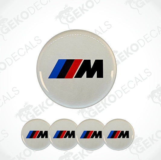 A set of four round car decals with a white background and a black 'M' logo with a red and blue stripe.
