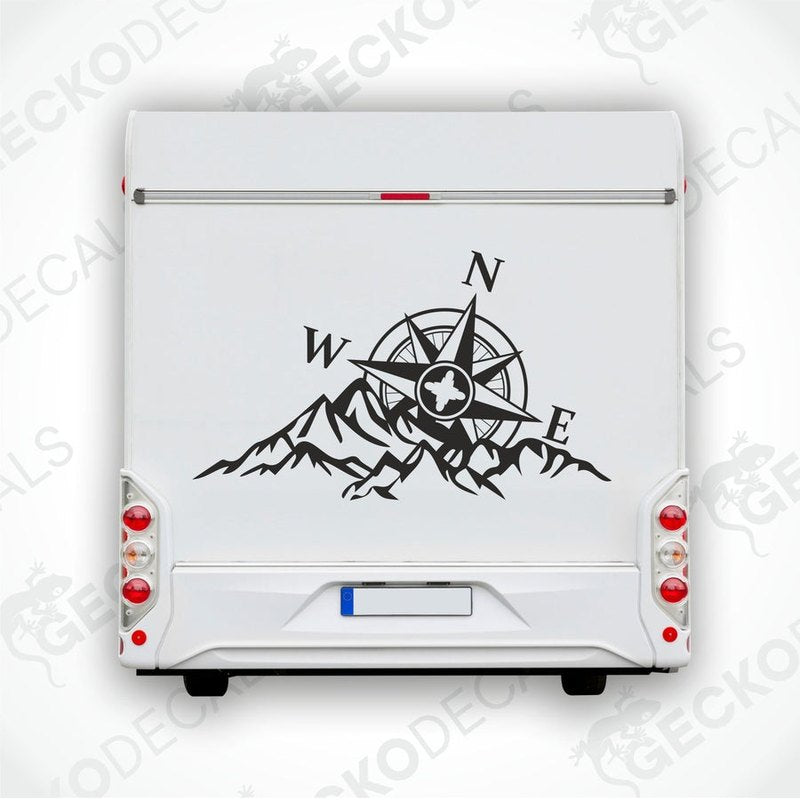 A white caravan with a black decal featuring a compass and mountains. The decal is positioned on the rear side of the caravan, near the window.