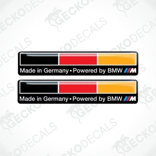 Two vinyl decals with 'MADE IN GERMANY POWERED BY BMW' text, one in black with a red border, and one in yellow with a black border.