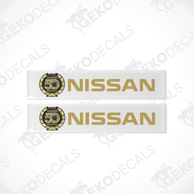 Two white vinyl stickers with gold and gold text and graphics representing the NISSAN brand, placed against a white background.