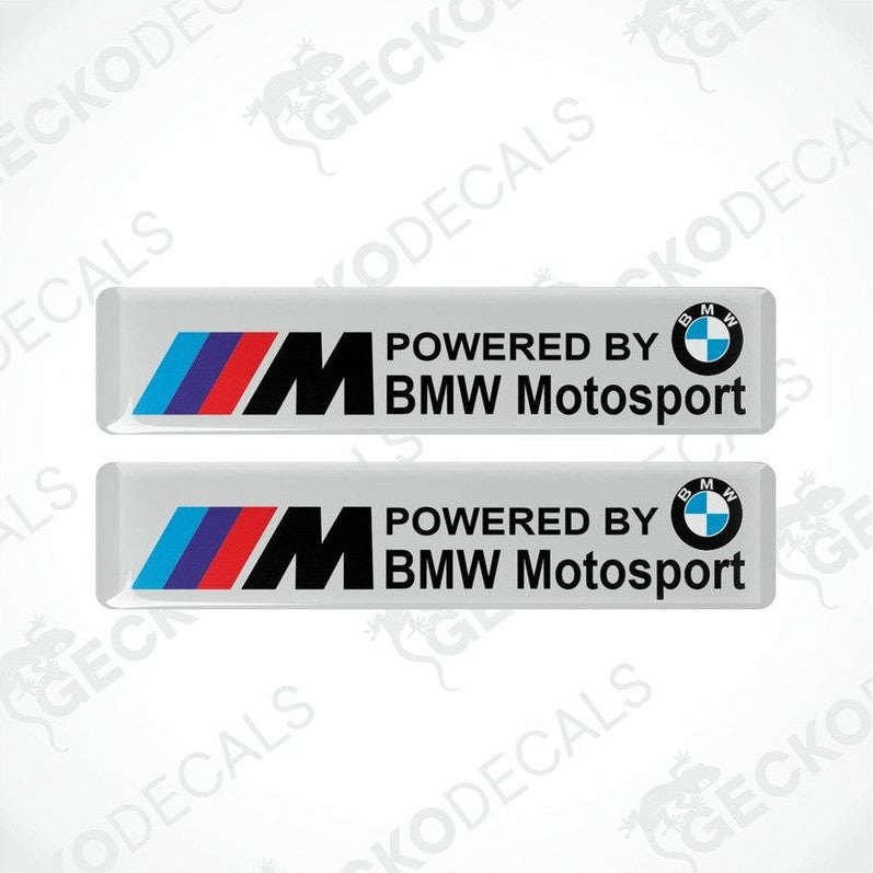 Two white vinyl stickers with the BMW Motosport logo, one featuring the BMW logo alone and the other with the text 'POWERED BY BMW MOTOSPORT M'. Both have a black, red, and white color scheme.