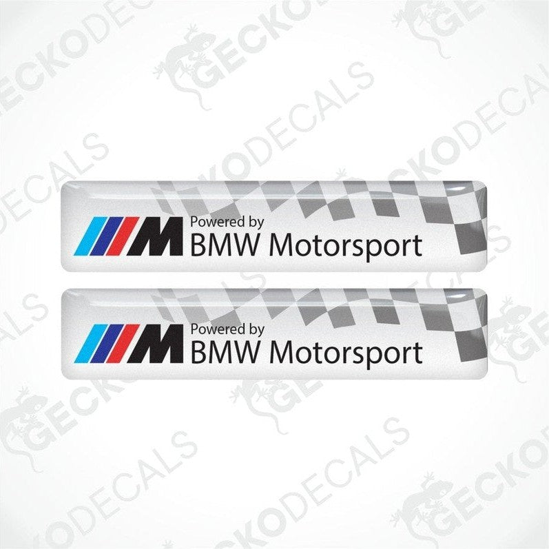 Two vinyl stickers with a  POWERED BY BMW M MOTORSPORT  logo, predominantly white with blue and red checkered pattern.