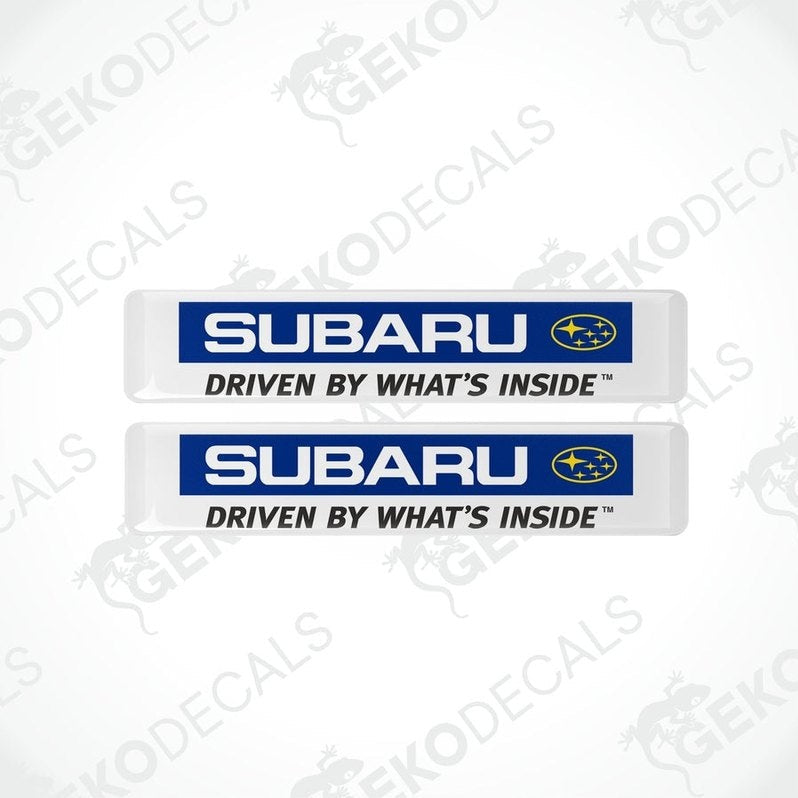 Two white vinyl stickers with the Subaru brand logo and the slogan 'DRIVEN BY WHAT'S INSIDE' printed in blue and yellow.