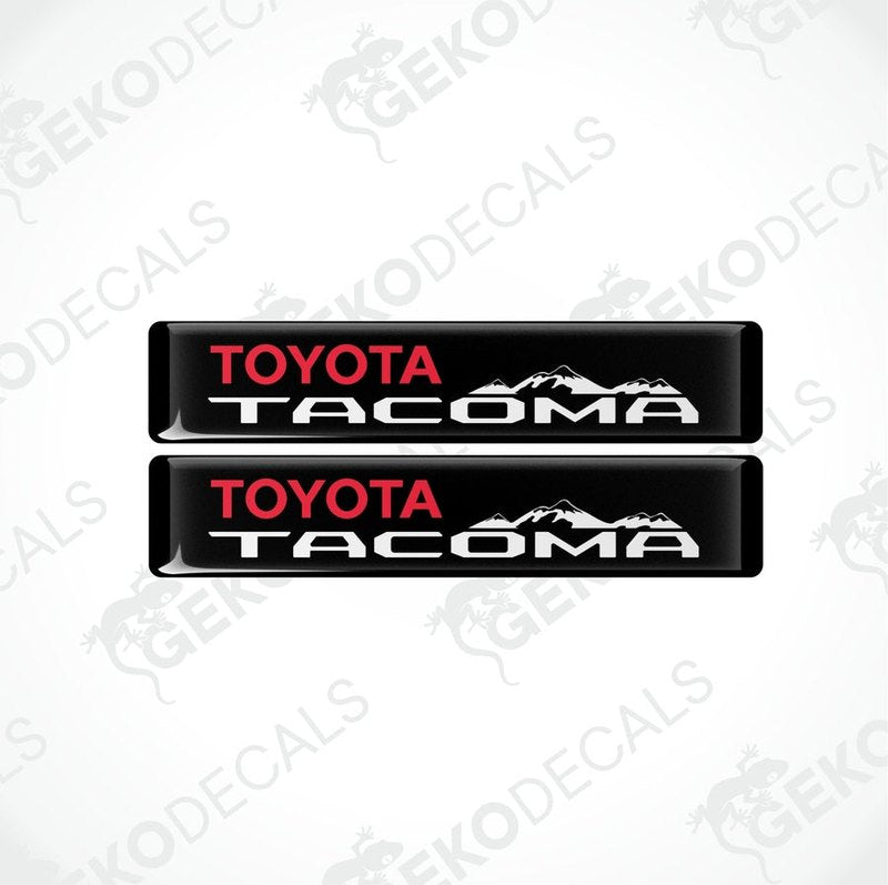 Two black vinyl decals with red and white text 'TOYOTA TACOMA' and a mountain range image.