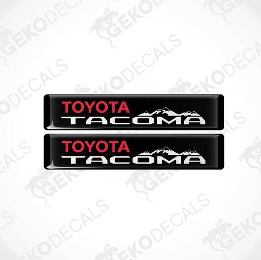 Two black vinyl decals with red and white text 'TOYOTA TACOMA' and a mountain range image.