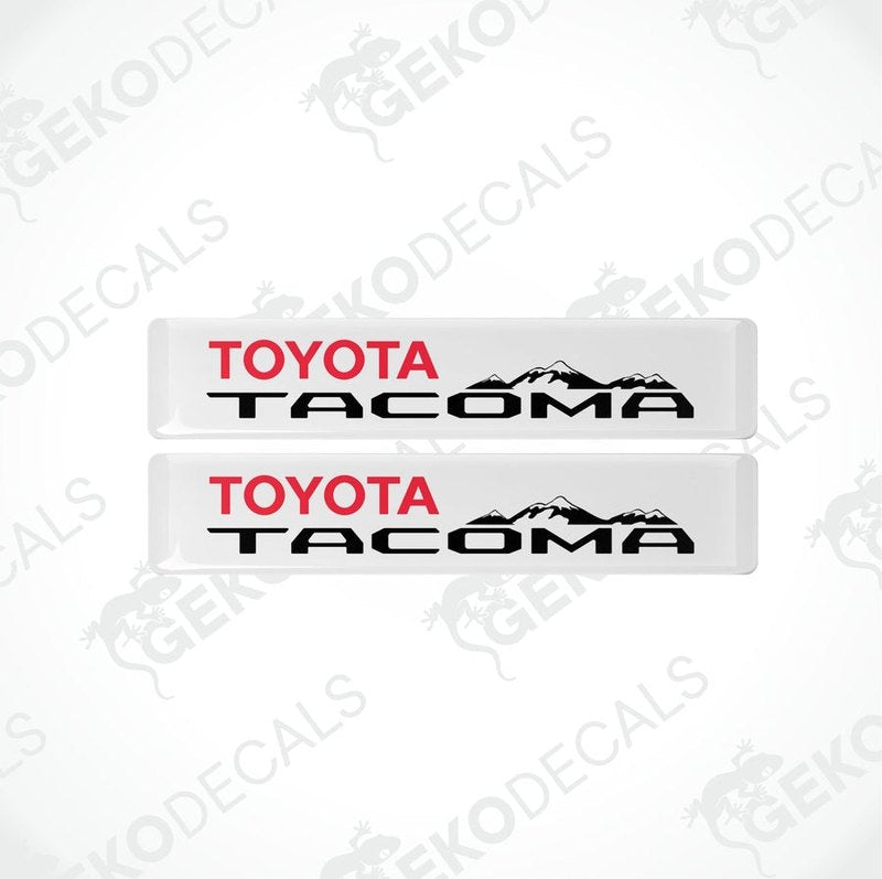Two withe vinyl decals with red and black text 'TOYOTA TACOMA' and a mountain range image.